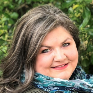 Outdoor headshot of a smiling Andrea Williams with wavy salt-and-pepper hair and a teal paisley scarf; soft natural light, leafy green background.