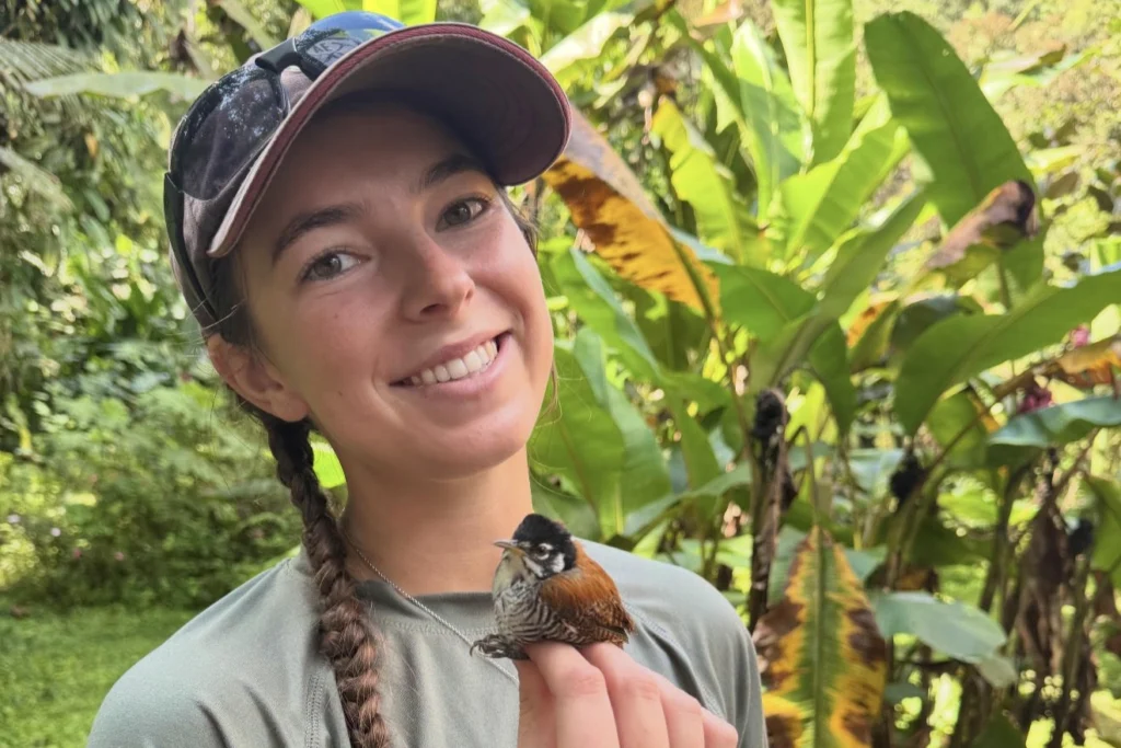 Hailey Rogan holds a small red, white and black bird while standing in dense forest in Ecuador