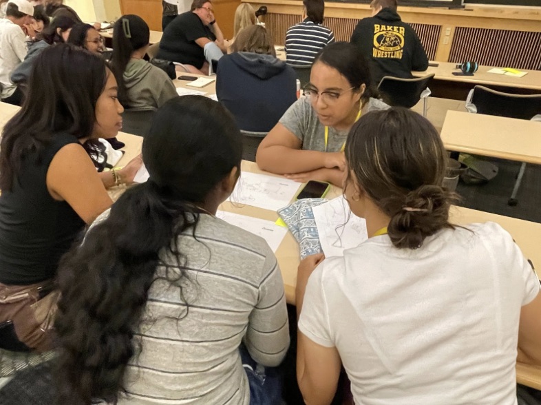 A group of students in a seminar sitting around a desk talking together 