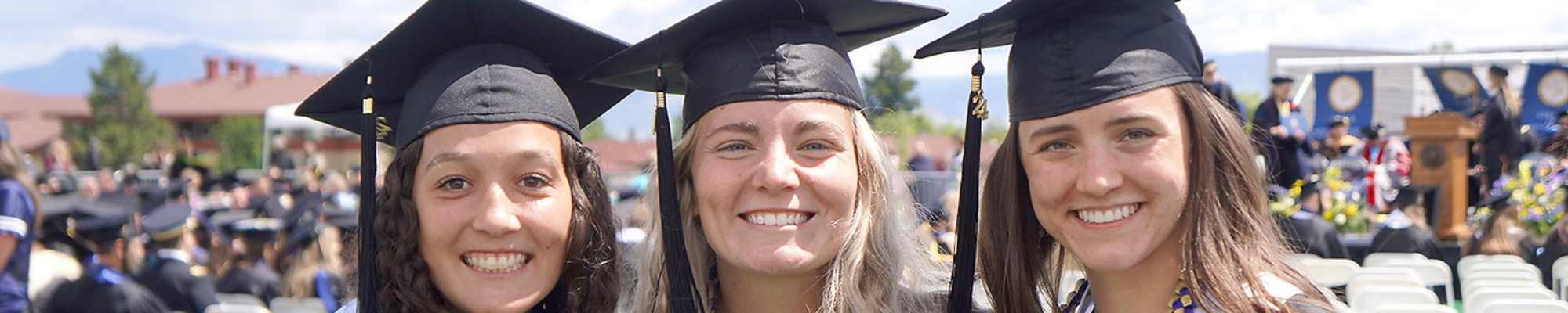 Three smiling graduates wearing black caps and gowns with white stoles pose together outdoors during a commencement ceremony. The background shows blurred spectators and campus buildings, suggesting a celebratory university setting.