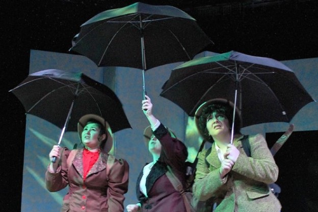 Three students in Victorian English costumes holding black umbrellas on stage and looking up