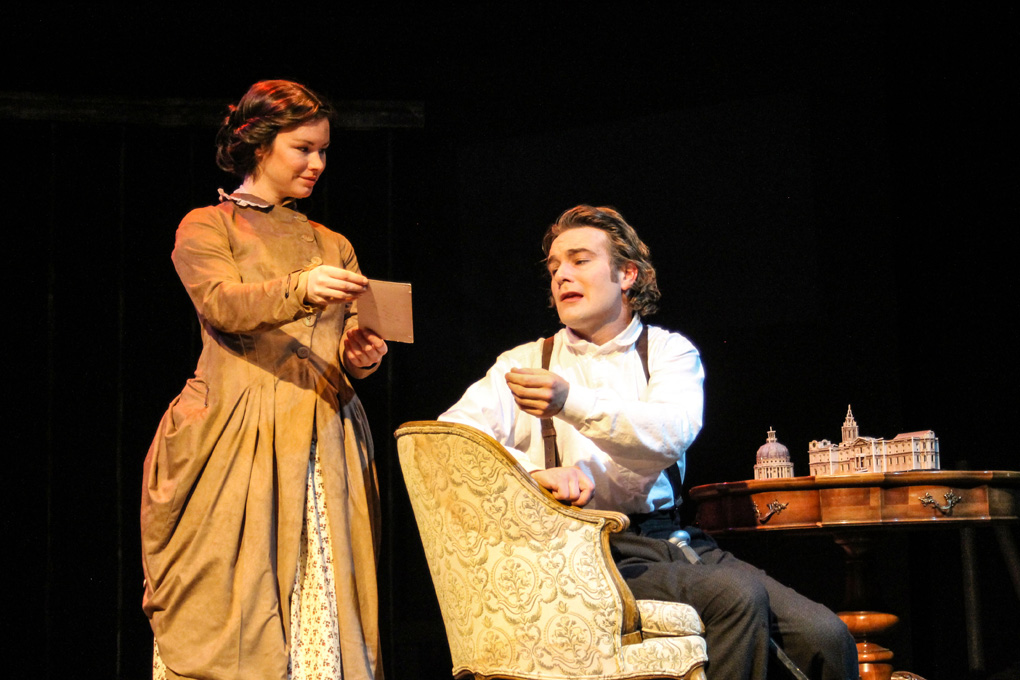 Two students in old fashioned formal wear on stage. A girl in a yellow dress holds out a note for a boy in a dress shirt and suspenders to read.
