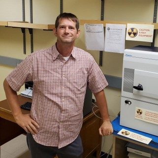 Man in short sleeved shirt standing next to a pieces of scientific equipment.