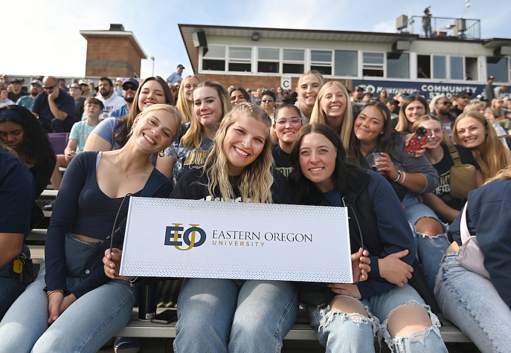 EOU Students sitting on the bleachers, smiling and holding an EOU placard