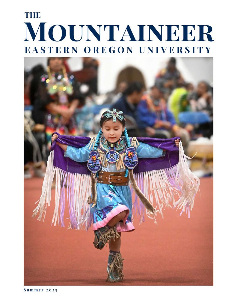 A young Native American girl dances gracefully in full regalia during a cultural event held at Eastern Oregon University. She wears a vibrant blue and purple traditional dress adorned with fringe, beadwork, and embroidered patches featuring floral and star designs. Her arms are extended outward, showcasing the flowing fringe of her outfit. The background is softly blurred, highlighting the child as the central focus, with spectators watching in the distance. The top of the image features the text "The Mountaineer, Eastern Oregon University" and the bottom reads "Summer 2025."
