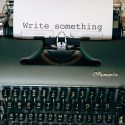 Close-up of a vintage Olympia typewriter with a sheet of paper that reads “Write something.”