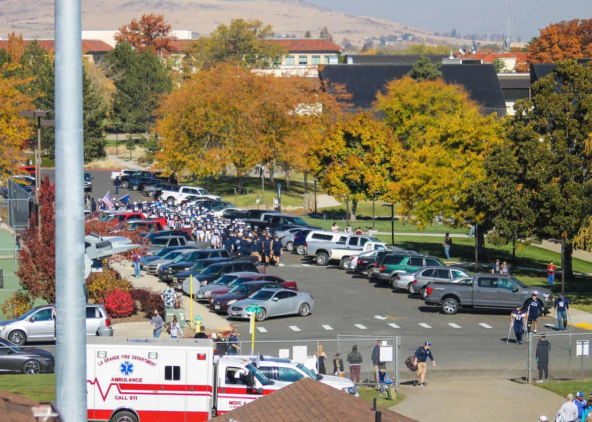 Elevated view of a sports team in dark blue uniforms marching through a college campus parking lot during an autumn homecoming event, lined with parked vehicles, vibrant fall foliage, and a La Grande Fire Department ambulance in the foreground.