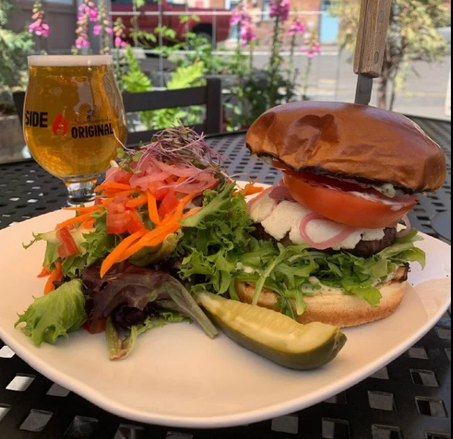 A gourmet burger held with a steak knife on a white plate next to a vibrant side salad, a pickle spear, and a glass of golden beer on an outdoor patio table.