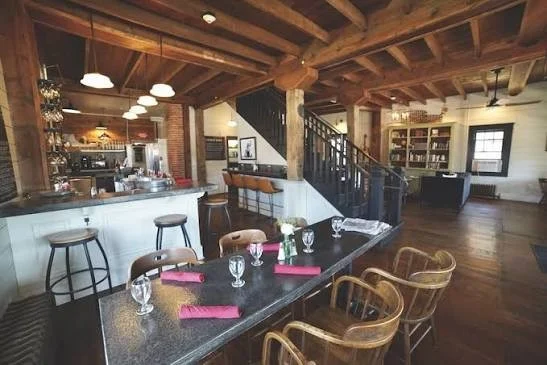 Cozy, rustic restaurant interior featuring exposed wood beam ceilings, a long dining table set with glasses and red napkins, a white bar counter with stools, and a central wooden staircase.